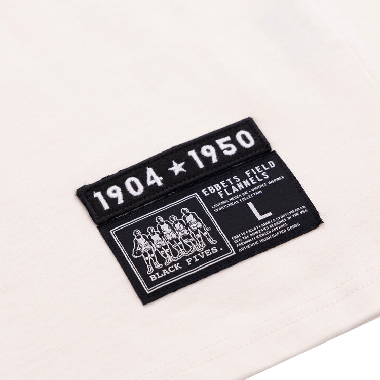 A close-up of the Black Fives T-Shirt - White by Ebbets Field Flannels, size L, featuring two black patches: one reads 1904 ★ 1950, the other highlights Black basketball history with BLACK FIVES and care details.