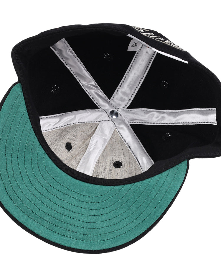 The image shows the inside of an Ebbets Field Flannels Black Fives Wool Ballcap, featuring a green underbrim, silver satin taping, center button, black ventilation holes, and a small white tag near the back.