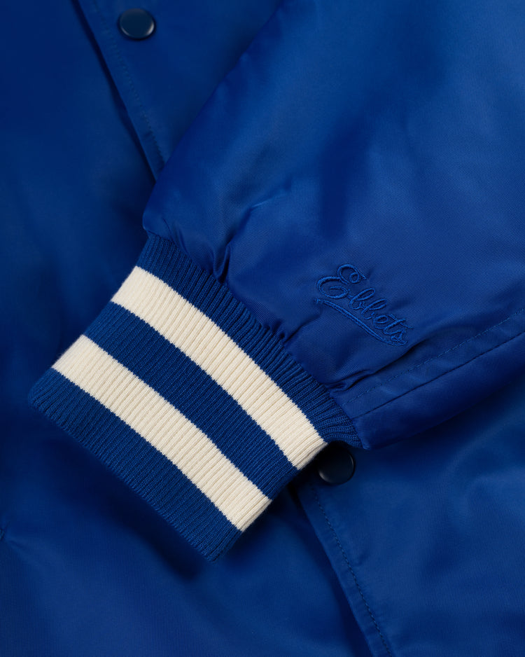 Close-up of a blue Los Angeles Angels (PCL) Satin Varsity Jacket sleeve by Ebbets Field, featuring white and blue striped ribbed cuff and Ebbets cursive embroidery near dark snap buttons.