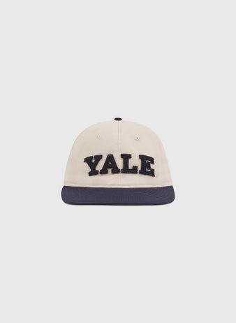 A cream-colored cotton twill ballcap by Ebbets Field, featuring “YALE” in bold chenille letters on the front and a dark blue brim, is displayed against a plain white background—perfect for showing your Yale University pride and timeless Ivy League style. The Yale Chenille Logo Cotton Twill Ballcap combines classic design with standout school spirit.