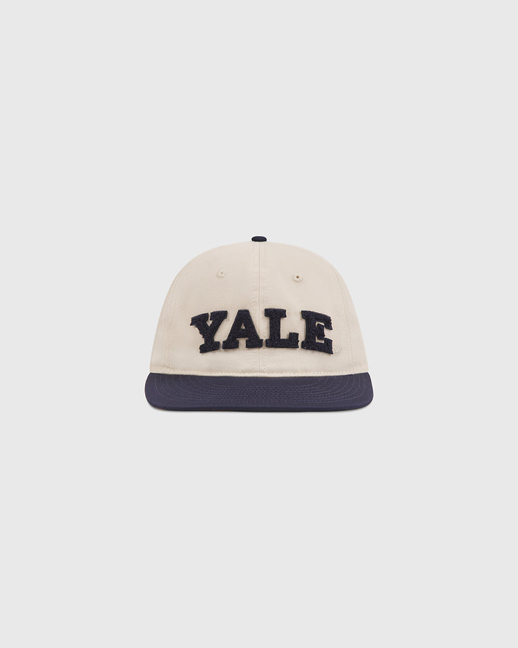 A cream-colored cotton twill ballcap by Ebbets Field, featuring “YALE” in bold chenille letters on the front and a dark blue brim, is displayed against a plain white background—perfect for showing your Yale University pride and timeless Ivy League style. The Yale Chenille Logo Cotton Twill Ballcap combines classic design with standout school spirit.