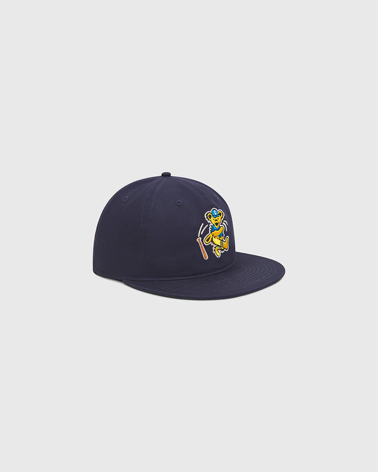 The Ebbets Field Grateful Dead Baseball Bear Cotton Twill Ballcap is a navy blue hat made from cotton twill, featuring a flat brim and an embroidered yellow and blue cartoon bear holding a bat on the front. The cap is displayed at an angle against a simple light background, evoking the playful style of classic Grateful Dead ballcaps.