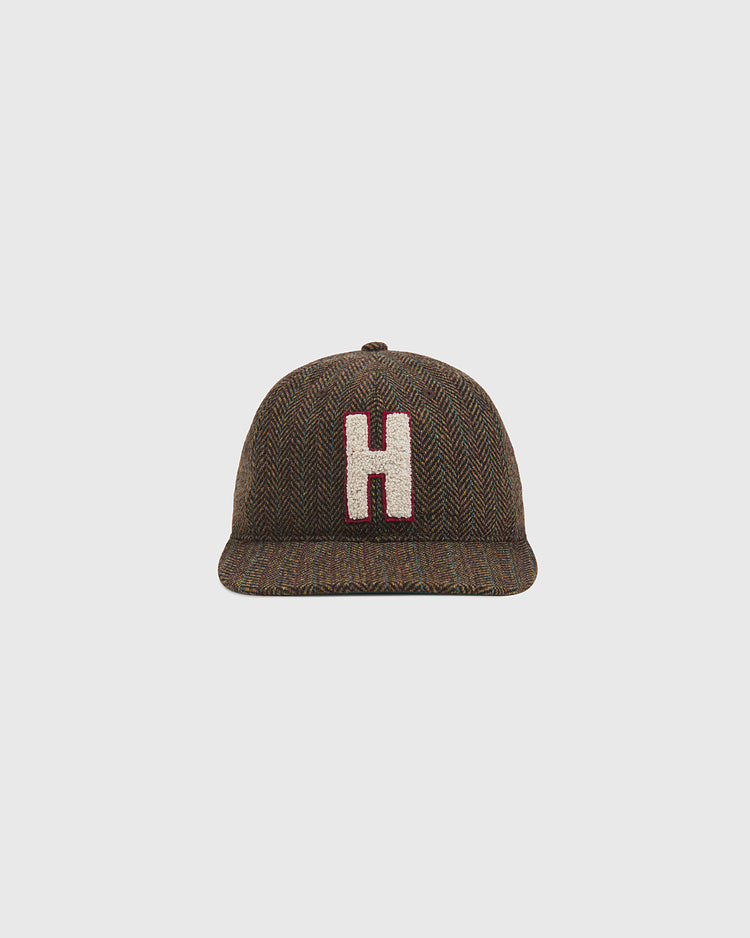 The Ebbets Field Harvard University Herringbone Wool Ballcap features a brown herringbone pattern, a flat brim, and a prominent white chenille patch letter “H” outlined in red on the front, shown against a plain light gray background.