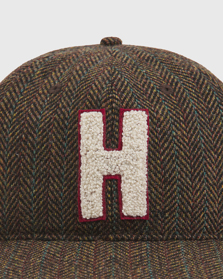 A close-up of the Harvard University Herringbone Wool Ballcap by Ebbets Field shows off its brown herringbone pattern and features a prominent, textured white chenille “H” outlined in red on the front—making it a standout accessory.