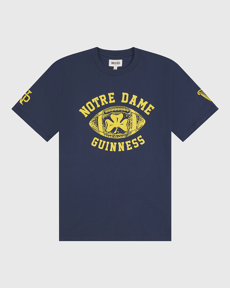 Show off your Irish heritage with the Ebbets Field Notre Dame X Guinness Short Sleeve Graphic Tee—a navy blue vintage shirt featuring bold yellow text, a football graphic with a shamrock, and striking yellow logos on both sleeves.