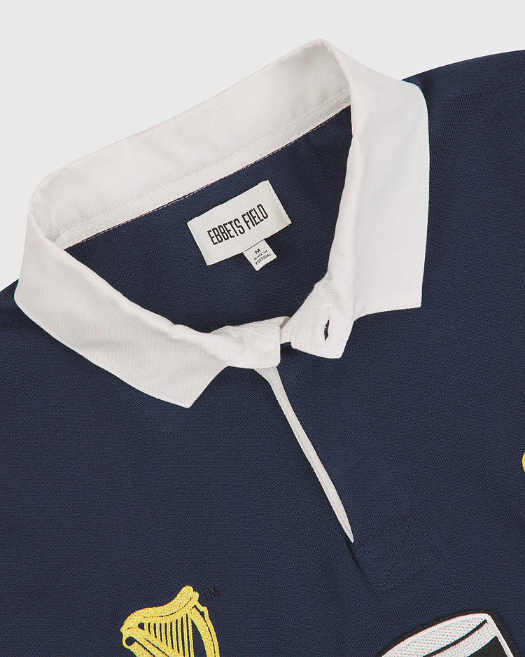 The Notre Dame X Guinness Rugby Shirt by Ebbets Field features a navy blue design with a white collar, drawing inspiration from classic Notre Dame rugby styling. Embroidered yellow and white patches evoke Irish heritage, while the Ebbets Field label is stitched inside.
