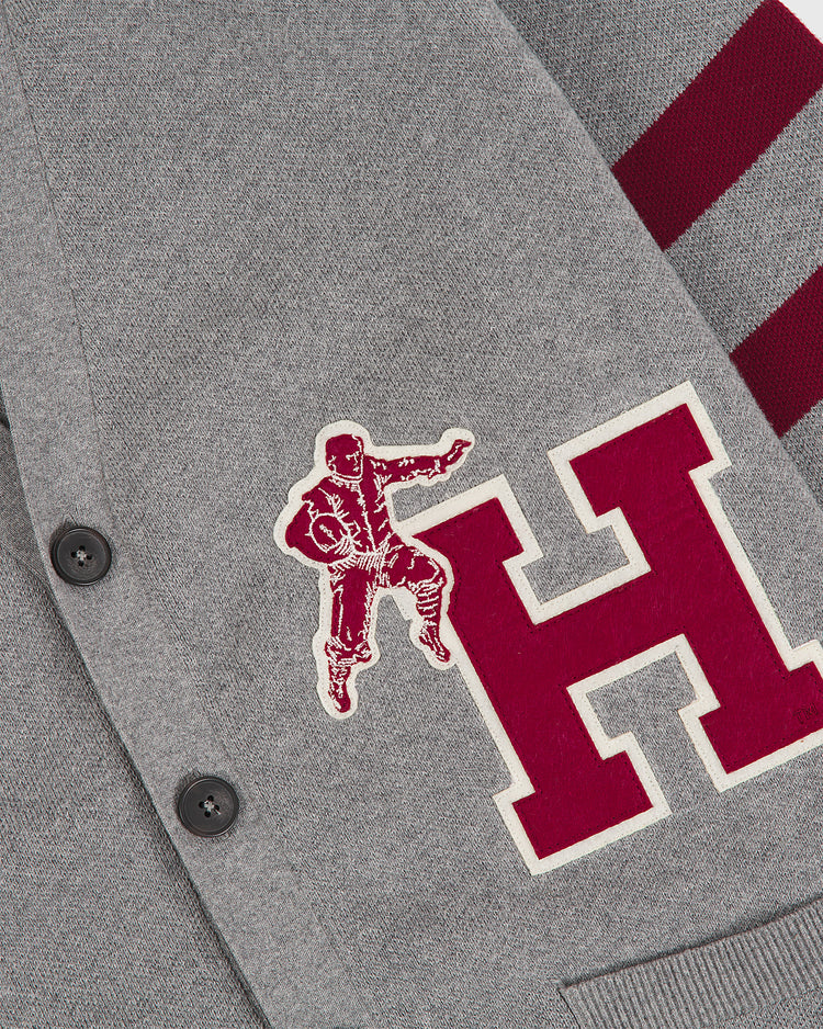 The Ebbets Field Harvard University Cotton Varsity Cardigan is a gray cardigan with black buttons, featuring a large red “H” and a vintage-style football player patch on the front, with two horizontal red stripes on the sleeve.