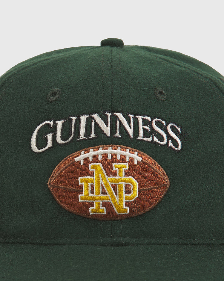 The Notre Dame X Guinness Wool Ballcap by Ebbets Field is a green wool cap featuring “GUINNESS” embroidered above a football with yellow interlocked ND letters, showcasing Notre Dame pride.