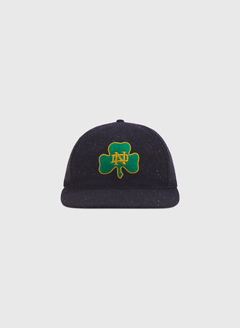 A Notre Dame X Magee Irish Donegal Wool Ballcap by Ebbets Field, in navy blue, features a green shamrock with yellow ND initials embroidered on the front and is displayed against a plain light gray background.