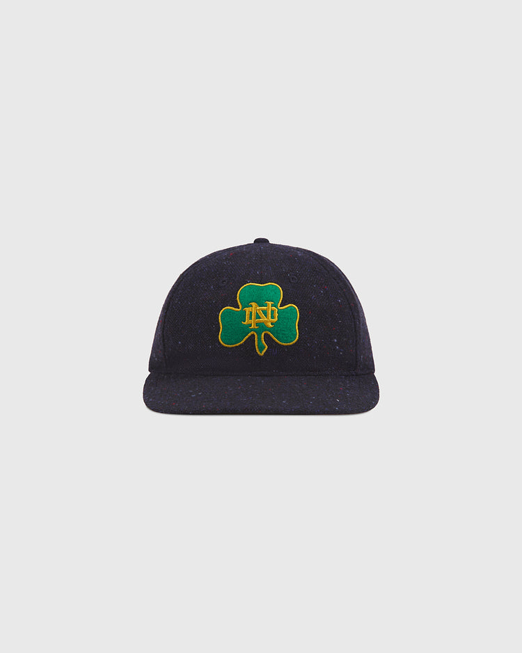A Notre Dame X Magee Irish Donegal Wool Ballcap by Ebbets Field, in navy blue, features a green shamrock with yellow ND initials embroidered on the front and is displayed against a plain light gray background.