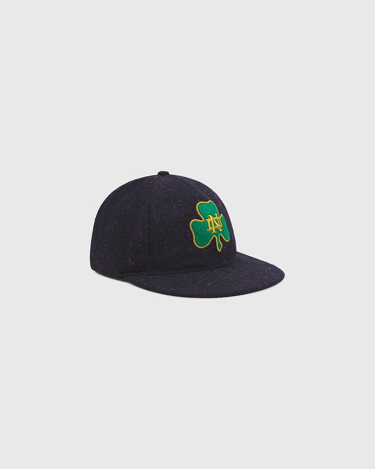 The Ebbets Field Notre Dame X Magee Irish Donegal Wool Ballcap, featuring a navy blue design with a green four-leaf clover and yellow ND lettering embroidered on the front, reminiscent of a classic Notre Dame hat, is displayed on a plain light gray background.