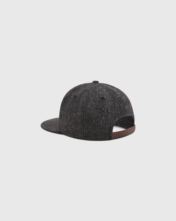 The Notre Dame X Magee Irish Donegal Wool Ballcap by Ebbets Field features a dark gray herringbone-patterned Donegal wool fabric with subtle colored specks. Shown from the back and side, this cap has a curved brim and an adjustable brown strap at the back.