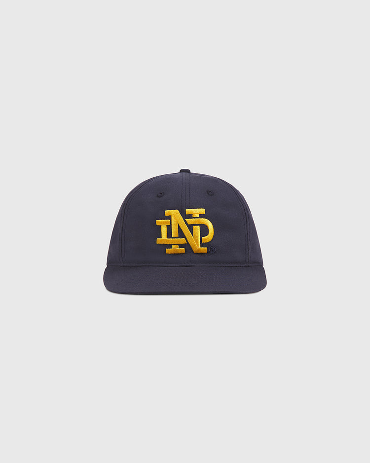 The Ebbets Field Notre Dame Cotton Ballcap is a navy blue baseball cap with a flat brim, featuring large yellow embroidered “NP” letters on the front, displayed on a plain light background.