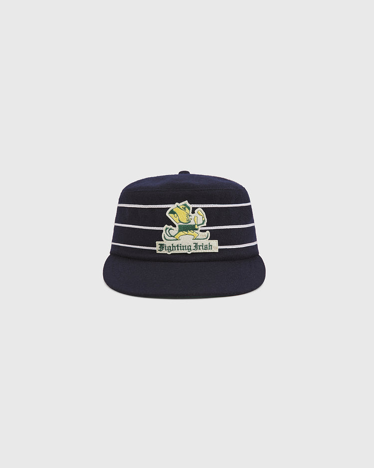 The Ebbets Field Notre Dame Pillbox Cap is a navy blue hat with white horizontal stripes and a leprechaun-adorned Fighting Irish logo on the front, displayed against a plain light gray background.