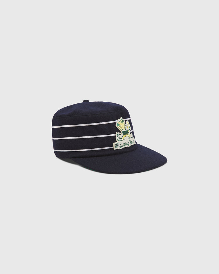 A Notre Dame Pillbox Cap by Ebbets Field, featuring navy blue fabric with white horizontal stripes and an embroidered green leprechaun logo above the words “Fighting Irish,” displayed against a plain light background.