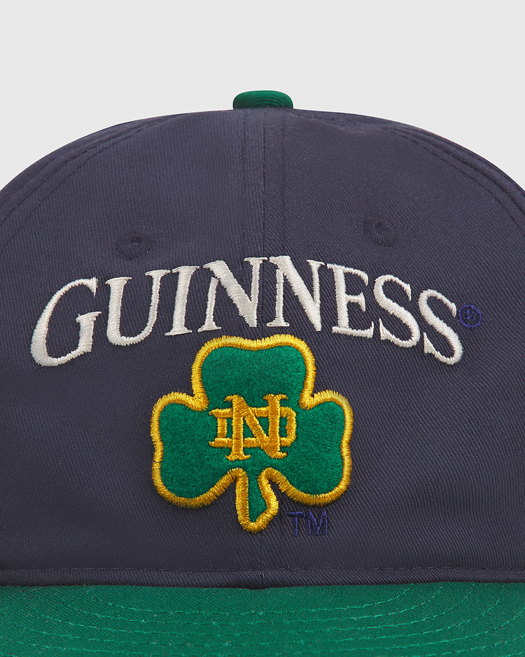 Introducing the Notre Dame X Guinness Cotton Ballcap by Ebbets Field—a navy hat featuring “GUINNESS” embroidered in white above a green shamrock with yellow ND initials, likely representing Notre Dame. The cap’s green brim adds an extra touch of Irish heritage, making this stylish ballcap a perfect blend of tradition and team spirit.