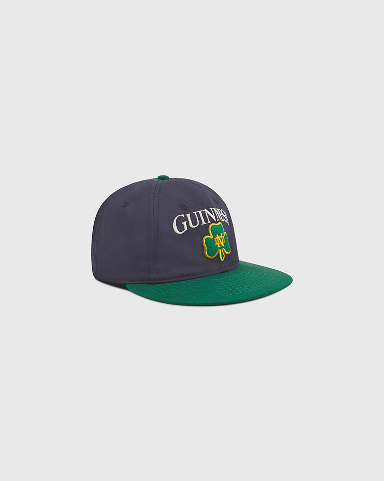 A navy blue and green Notre Dame X Guinness Cotton Ballcap by Ebbets Field, featuring GUINNESS and a yellow-outlined green shamrock embroidered on the front—perfect for celebrating your Irish heritage.