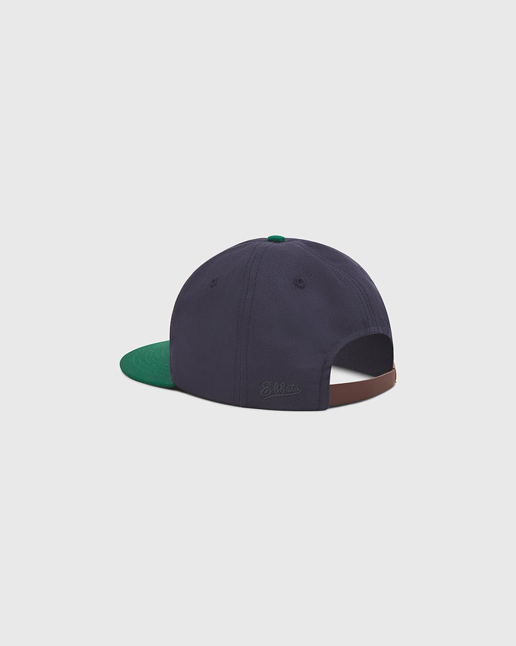 A navy blue Notre Dame X Guinness cotton ballcap by Ebbets Field, featuring a green bill and brown accent, is shown from the back with an opening and subtle embroidered text above it—this premium hat celebrates Irish heritage with understated style.