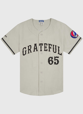 Introducing the Grateful Dead Cotton Twill Button-Up Baseball Jersey by Ebbets Field—a beige cotton twill jersey styled after iconic Grateful Dead apparel, with “GRATEFUL” and the number 65 boldly printed in black on the front. Finished with a red, white, and blue circular patch on the left sleeve, this piece is perfect for fans of psychedelic band fashion.