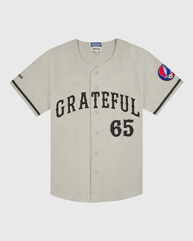 Introducing the Grateful Dead Cotton Twill Button-Up Baseball Jersey by Ebbets Field—a beige cotton twill jersey styled after iconic Grateful Dead apparel, with “GRATEFUL” and the number 65 boldly printed in black on the front. Finished with a red, white, and blue circular patch on the left sleeve, this piece is perfect for fans of psychedelic band fashion.