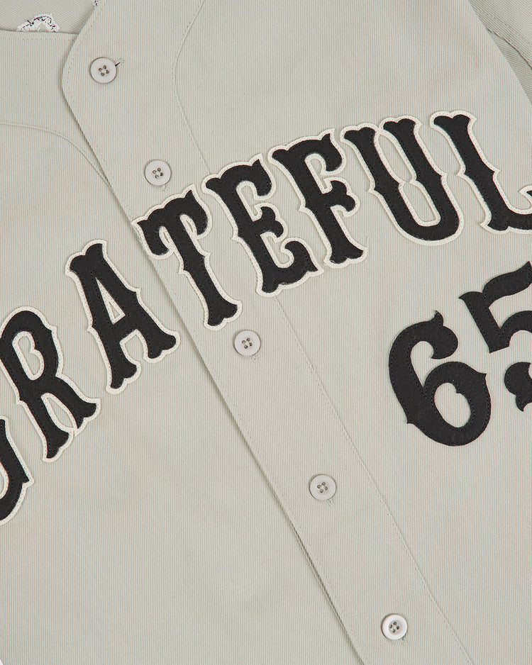 A close-up of the Ebbets Field Grateful Dead Cotton Twill Button-Up Baseball Jersey, featuring light-colored cotton twill fabric with “GRATEFUL” in bold black letters and the partially visible number 65 on the right side.