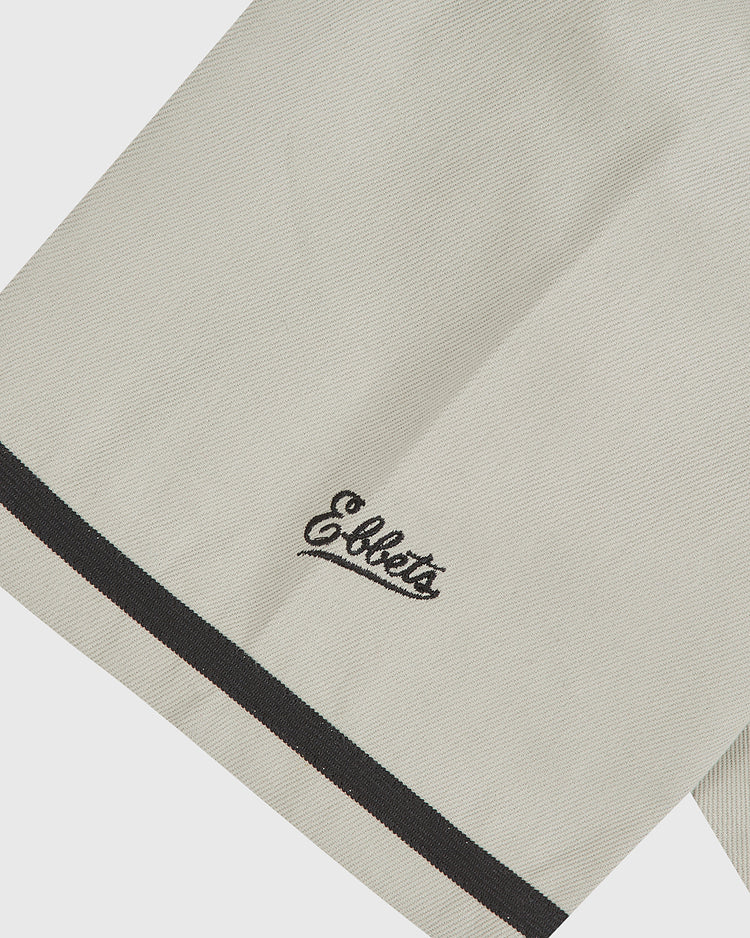 A close-up of the Grateful Dead Cotton Twill Button-Up Baseball Jersey by Ebbets Field, featuring beige cotton twill fabric with a black border and the word “Ebbets” embroidered in black script near the edge, evoking vintage psychedelic band style.