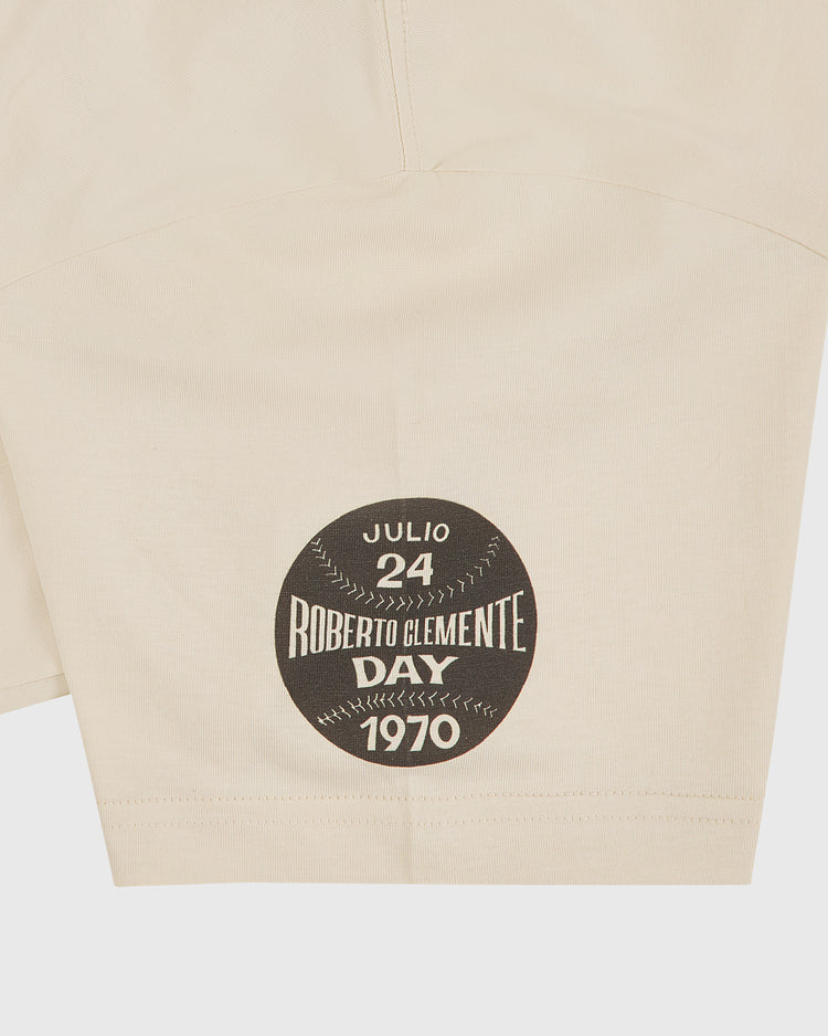 The Clemente Museum x Ebbets Field Short Sleeve Graphic T-Shirt by Ebbets Field features beige fabric with a black circular patch and white text reading “Julio 24 Roberto Clemente Day 1970,” designed to resemble a baseball—an ideal tribute for Pittsburgh Pirates shirt collectors or fans of classic Roberto Clemente graphic tees.