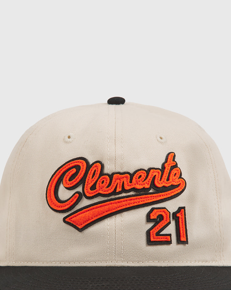 The Ebbets Field Clemente Museum X Ebbets Field Cotton Twill Ballcap is a beige and black cap made from cotton twill, featuring custom Clemente script and the number 21 embroidered in orange and black on the front, inspired by the Roberto Clemente Museum.