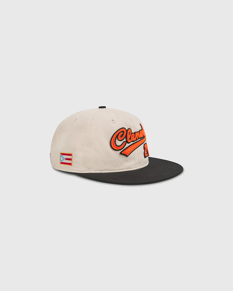 The Clemente Museum X Ebbets Field Cotton Twill Ballcap by Ebbets Field features a cream and black cotton twill construction with custom Clemente script in orange, a small Puerto Rican flag patch on the side, and a flat black brim—created in collaboration with the Roberto Clemente Museum.