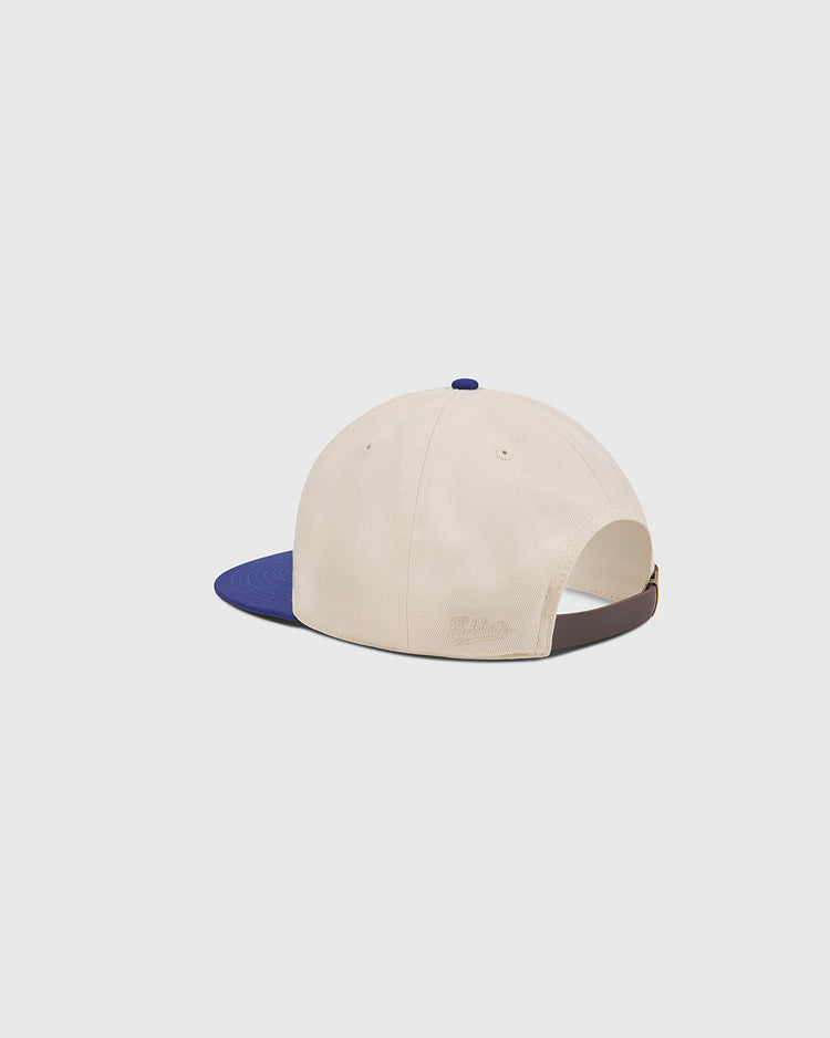 The Ebbets Field Hollywood Stars 1957 Cotton Twill Ballcap is shown from the back, featuring a beige cotton twill body with a contrasting blue brim, an adjustable brown strap, and a small blue button on top. The classic design is inspired by the timeless style of the 1957 Hollywood Stars at Gilmore Field. The background is plain white.