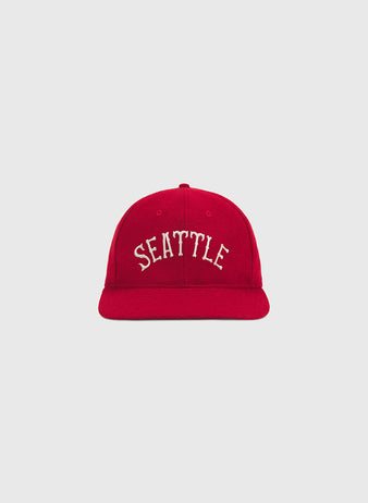 The Seattle City Series Wool Ballcap by Ebbets Field features a vibrant red design with “SEATTLE” embroidered in white capital letters on the front, expertly crafted from genuine wool broadcloth and shown against a plain light gray background—an essential piece for any city series hat collection.