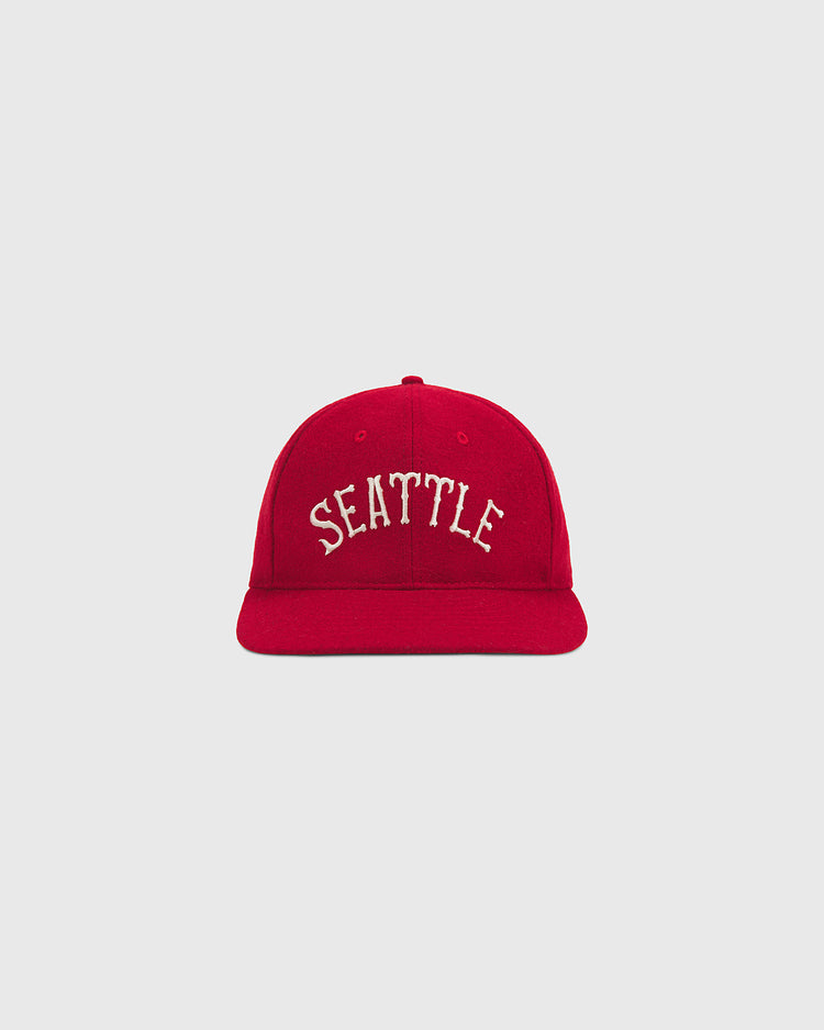 The Seattle City Series Wool Ballcap by Ebbets Field features a vibrant red design with “SEATTLE” embroidered in white capital letters on the front, expertly crafted from genuine wool broadcloth and shown against a plain light gray background—an essential piece for any city series hat collection.