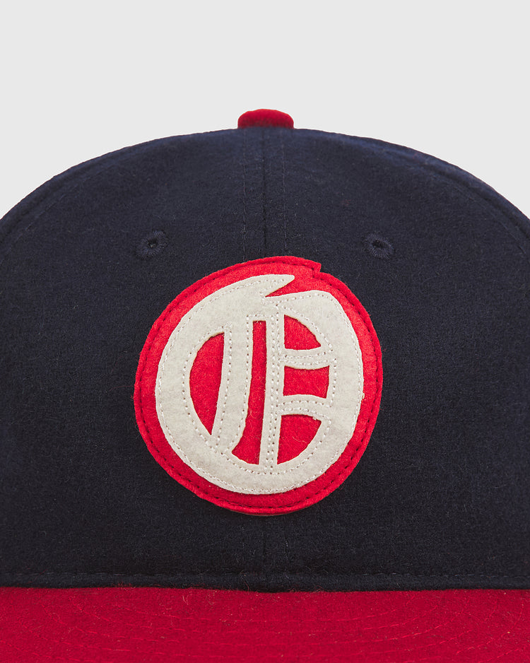 A navy blue and red Ebbets Field Oakland Oaks 1948 Authentic Wool Ballcap, featuring a white and red circular embroidered logo with stylized lettering on the front, reminiscent of a classic vintage baseball hat.