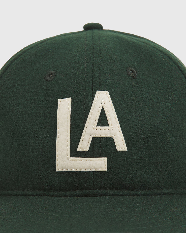 An authentic dark green wool ballcap by Ebbets Field, featuring large white “LA” embroidery on the front and inspired by the 1942 Los Angeles Angels of the PCL, is shown in a front-facing photograph against a plain background.