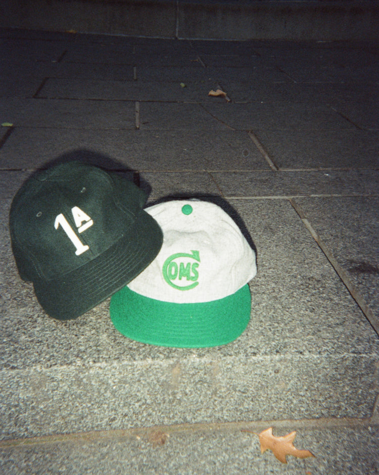 Two Mystery Caps of Mexico – Artelleria de México 1926 Wool Ballcaps rest on stone steps; one is dark green with 1A on the front, and the other is light gray with a green brim and a circular OMS logo.