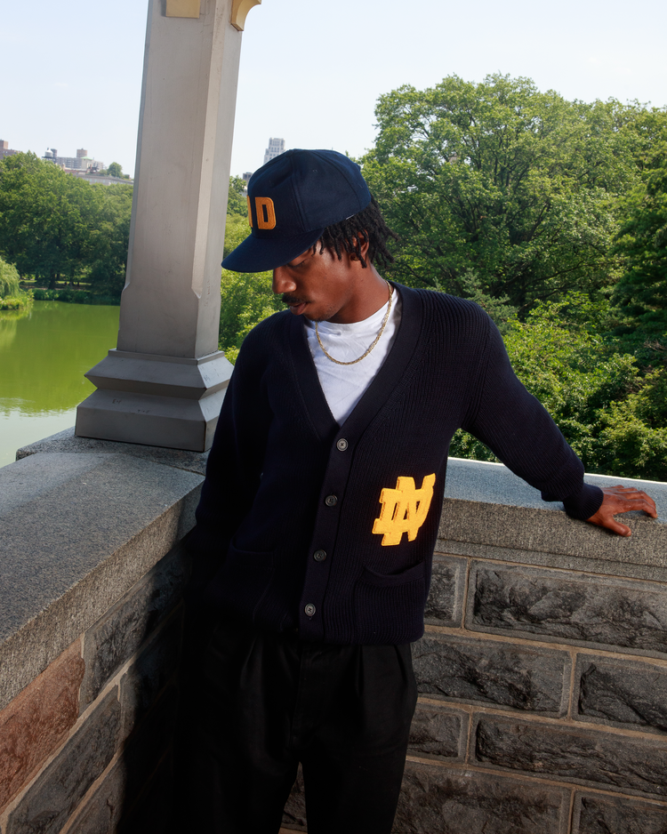 A young man wearing the Ebbets Field University of Notre Dame Varsity Cardigan and a matching cap stands on a stone balcony overlooking greenery and a lake, gazing downward with one hand resting on the ledge.