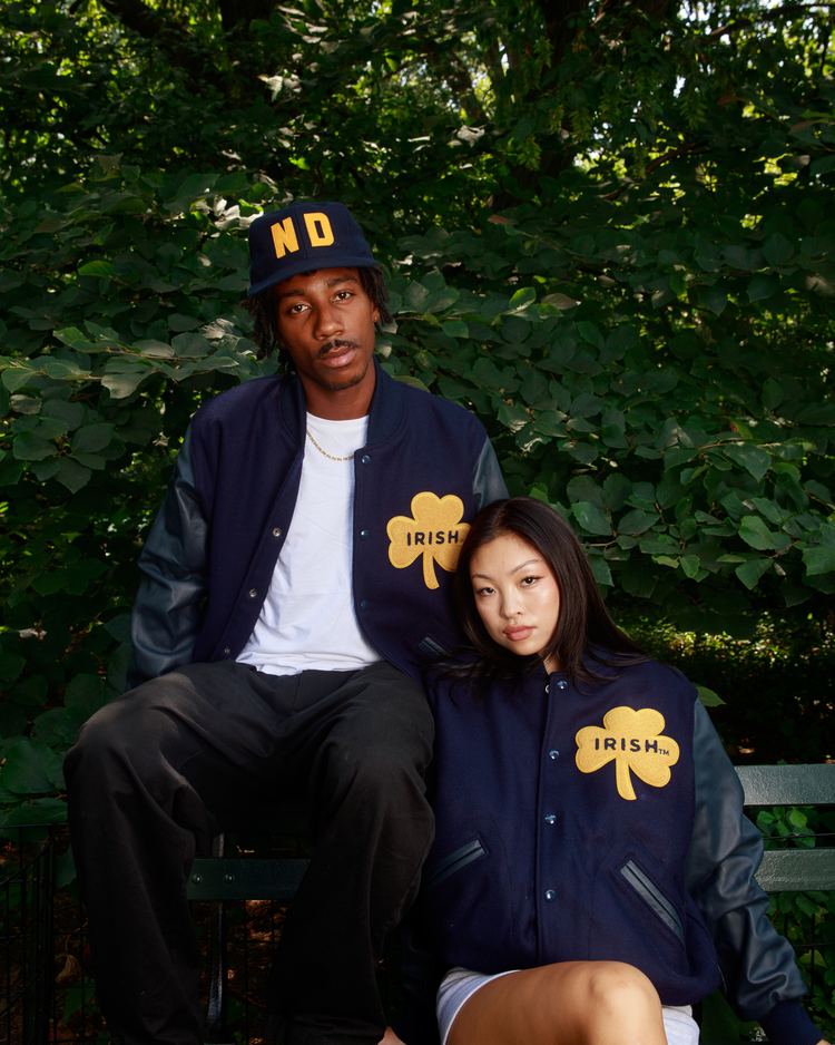 Two young adults sit outside in matching EFF University of Notre Dame Authentic Wool & Leather Varsity Jackets with large yellow shamrocks and IRISH lettering. One wears a navy ND cap, surrounded by lush green foliage.