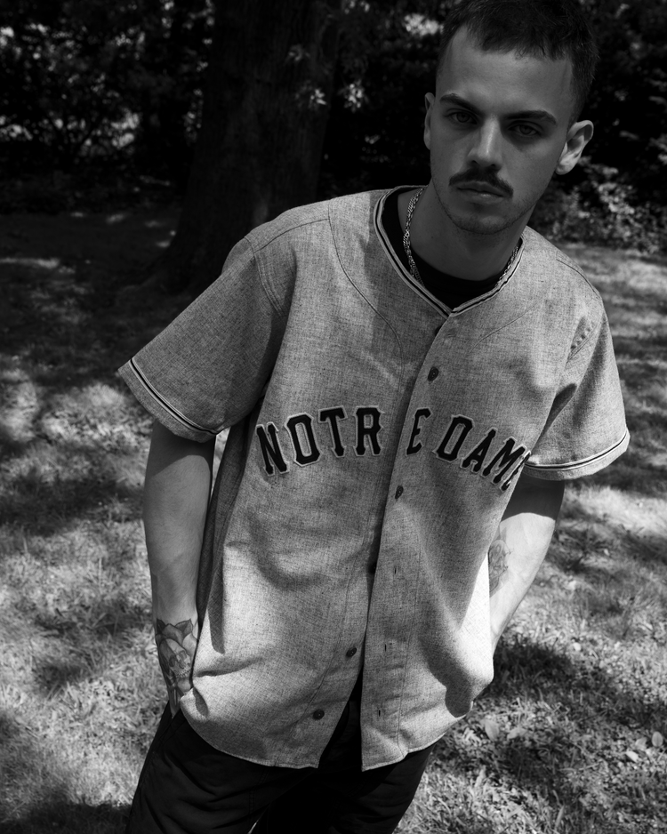 A man with short hair and a mustache stands outdoors in partial shade, wearing an Ebbets Field University of Notre Dame 1956 Authentic Wool Road Baseball Jersey and dark pants. One hand in his pocket, he gazes seriously at the camera.