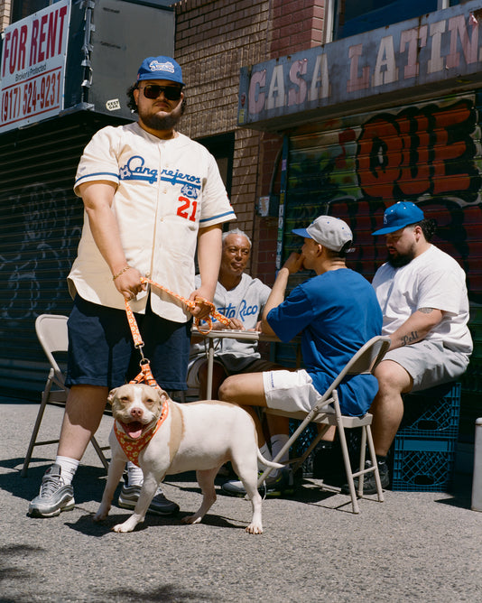 A man in a Dodgers jersey and cap stands on a sunny street holding a leashed dog, while four people sit around a table behind him. A “Casa Latina” sign and a “For Rent” sign are visible in the background.