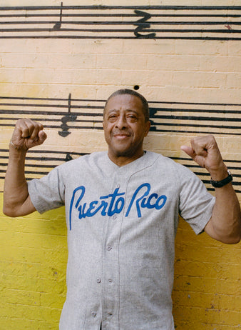 An older man wearing a Puerto Rico baseball shirt stands in front of a yellow wall with black musical notes, flexing both arms and smiling confidently at the camera.