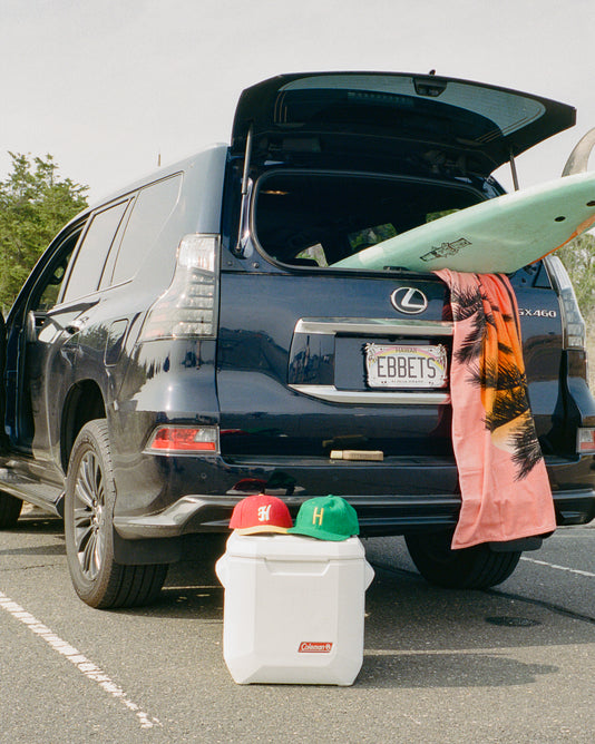 A black Lexus SUV with its trunk open shows a surfboard and towel inside. Two baseball caps rest on a white cooler behind the car in a sunny parking lot.