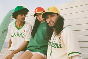 Three people wearing Hawaii-themed baseball shirts and caps sit by a white wooden wall at the beach, with a green surfboard and the ocean in the background. They look relaxed and casual.