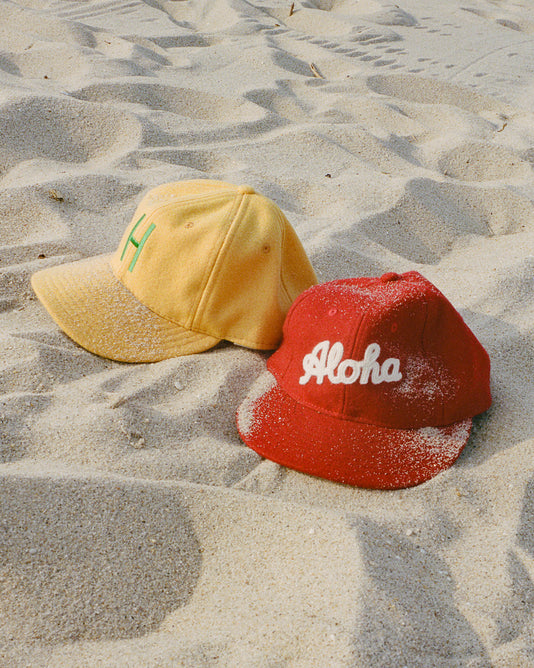 Two baseball caps on sandy beach: one yellow cap with a green H and one red cap with white Aloha text, both partially covered in sand.