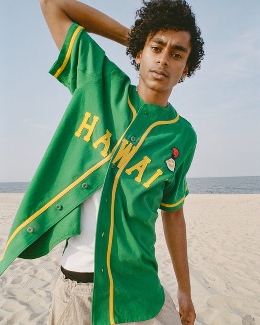 A young person wearing a green HAWAII baseball jersey, white shirt, and beige shorts stands on a sandy beach, holding one arm behind their head, with the ocean and blue sky in the background.