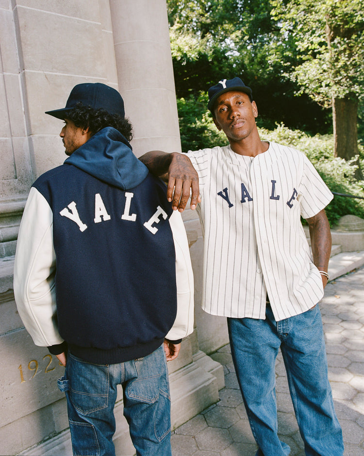 Two men stand outside a stone building in Yale-branded attire. One faces away, wearing the Ebbets Field Yale University Wool & Leather Varsity Jacket and cap, while the other in a 1950s-inspired Yale baseball jersey leans against him amid trees.