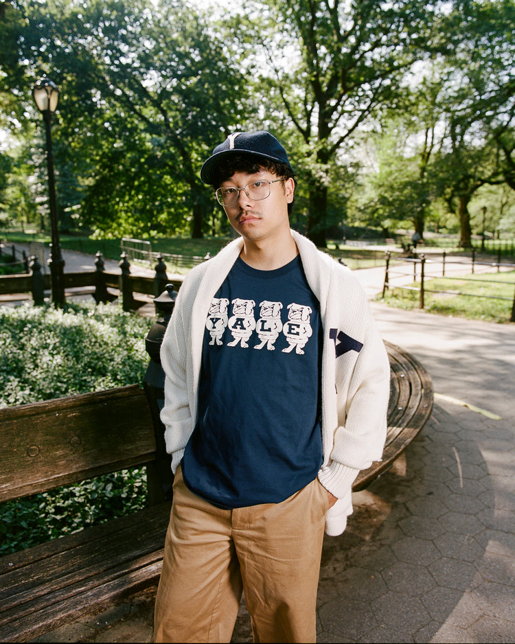 A young man in an Ebbets Field Yale University Short Sleeve Tee, light cardigan, cap, and glasses stands by a wooden bench in a sunlit park with green trees.