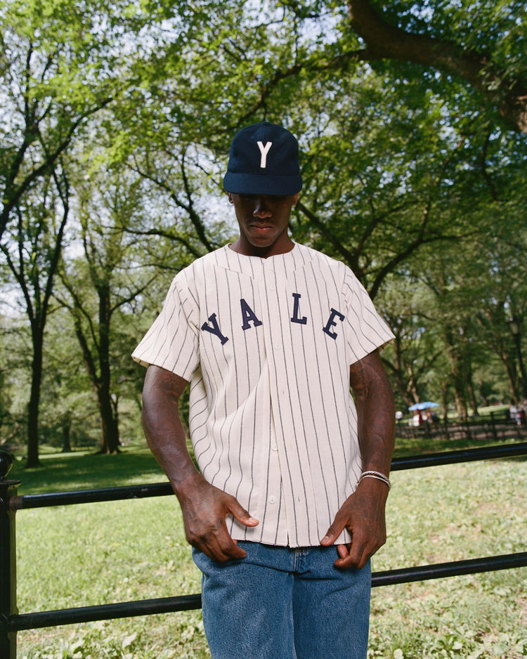 A person wearing an Ebbets Field Yale University 1948 Authentic Wool Home Baseball Jersey and a navy Y cap stands outdoors, with trees and a black metal fence in the background.