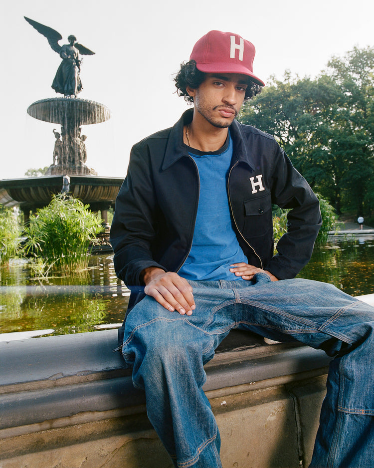 A young man in an Ebbets Field Harvard University Cotton Grounds Crew Jacket and a matching red Harvard cap sits by a fountain in a park, with a winged statue and lush greenery in the background.