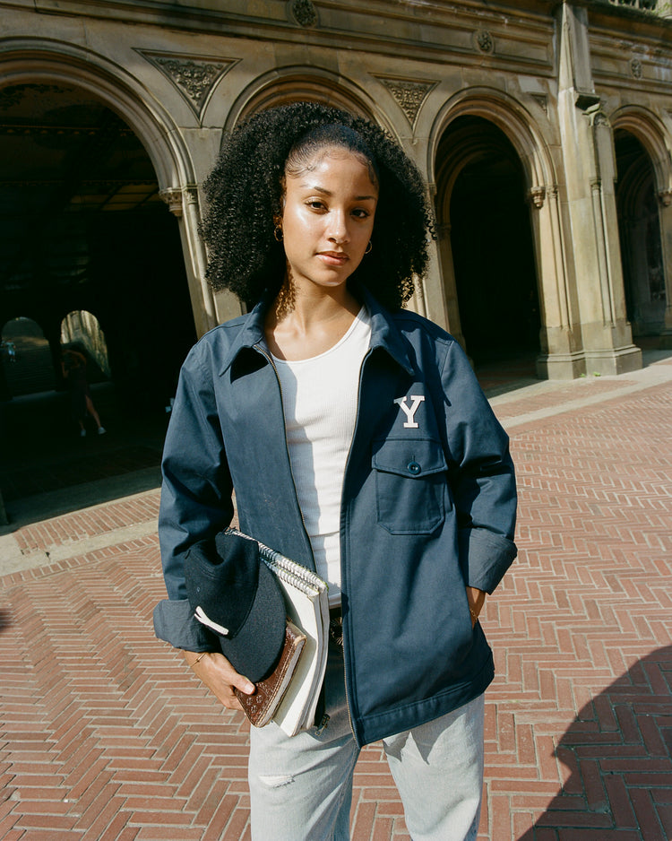 A person with curly hair stands outdoors before arched stone architecture, wearing an Ebbets Field Yale University Cotton Grounds Crew Jacket with a Y on the chest, and holding notebooks and a cap.
