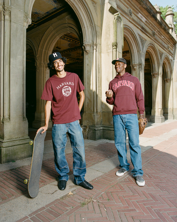 Two men stand outside a historic stone building in Harvard shirts and jeans. One, holding a skateboard, smiles; the other gestures with a baseball glove and wears an Ebbets Field Harvard University 1950 Authentic Wool Ballcap.